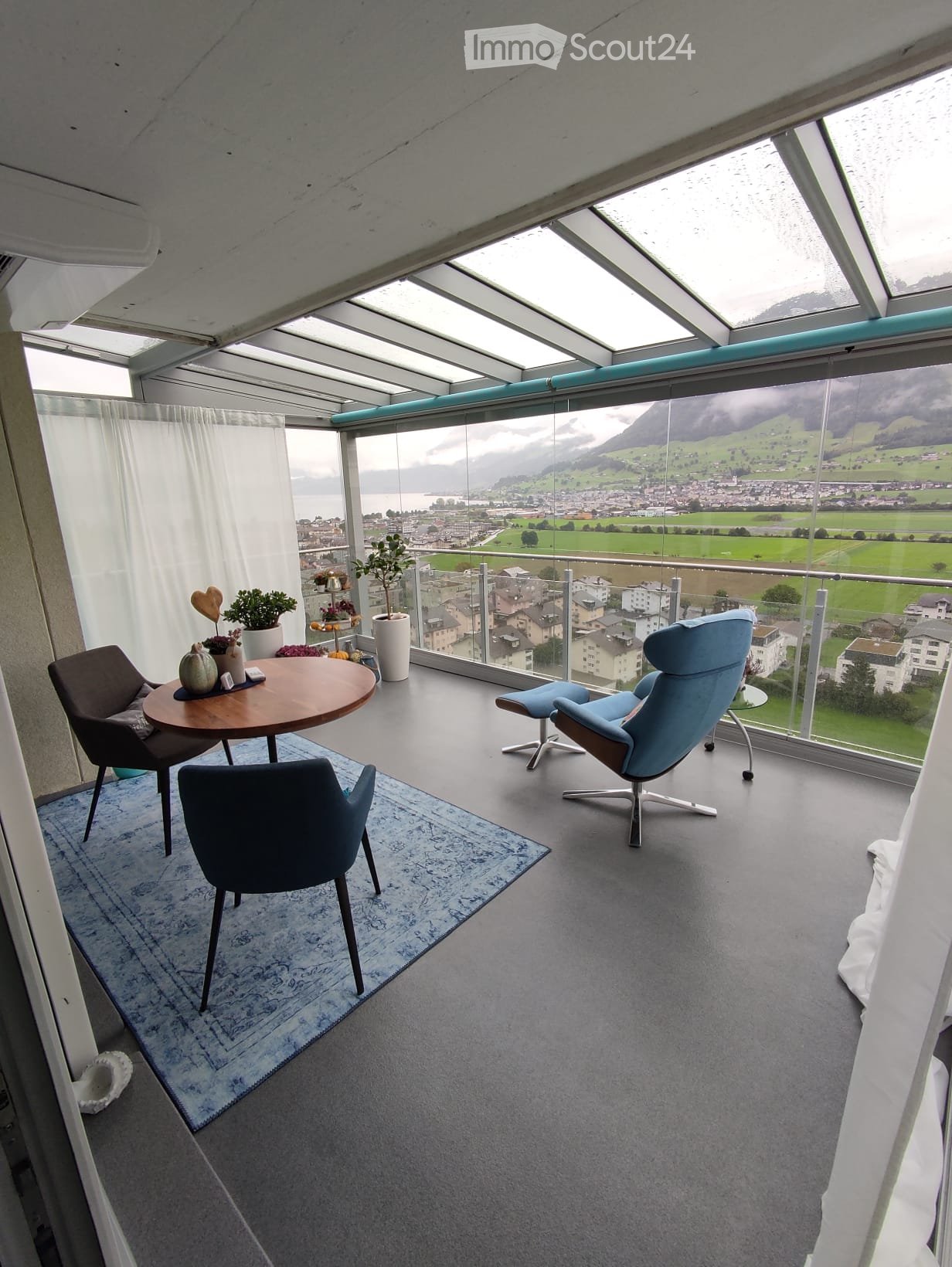 Modern balcony with panoramic mountain view, glass ceiling, grey carpet, blue rug, round wooden table, chairs, and potted plants.