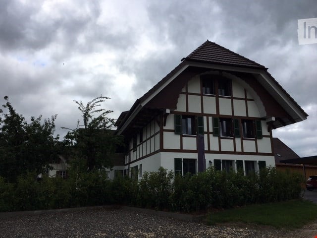 Traditional Swiss-style house with white walls, dark wooden trim, and a steeply pitched roof. The house features multiple windows and is surrounded by greenery and a gravel driveway.