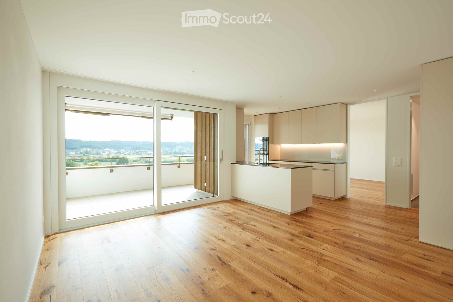 Modern, minimalist kitchen with light wood flooring, white cabinets, and a central island. Large sliding glass doors open to a balcony with scenic views.