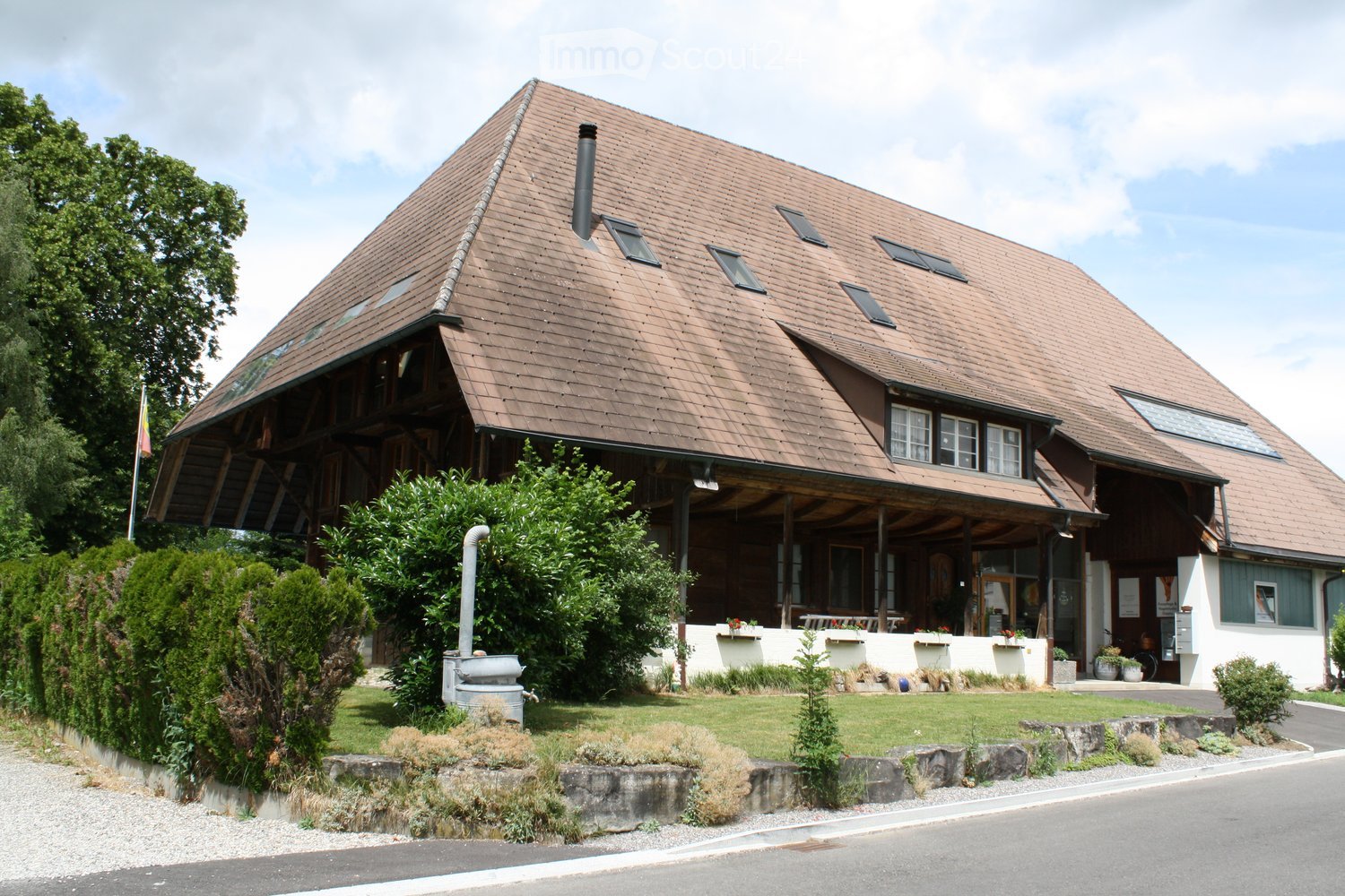A two-story wooden house with a steep, brown tiled roof and multiple dormer windows. The house features a covered porch area with seating and flower boxes, surrounded by a well-maintained garden with shrubs and trees.