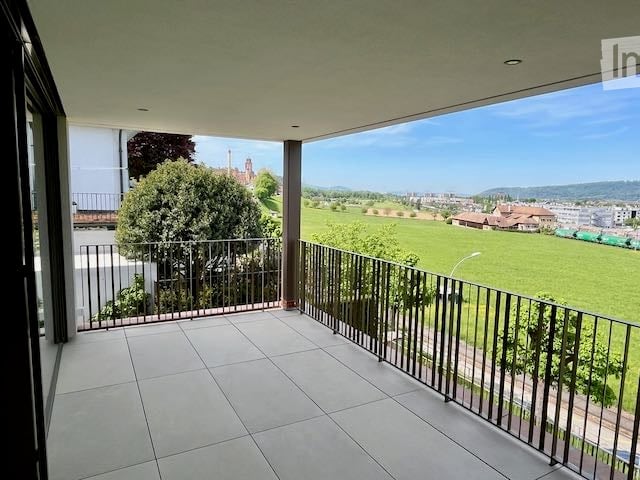 Spacious balcony with tiled floor, metal railing, and scenic view of a green field and town.