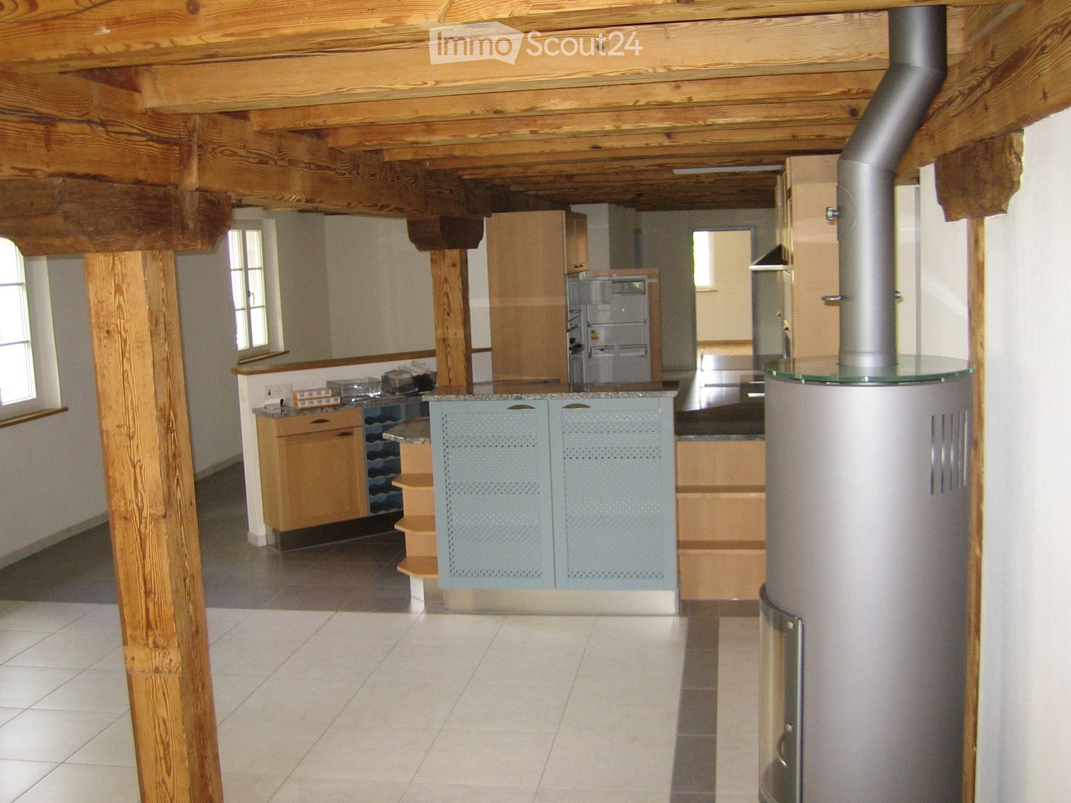 Open-plan kitchen and living area with exposed wooden beams, light-colored cabinets, a refrigerator, and a stove. The space features a tiled floor and large windows allowing natural light.