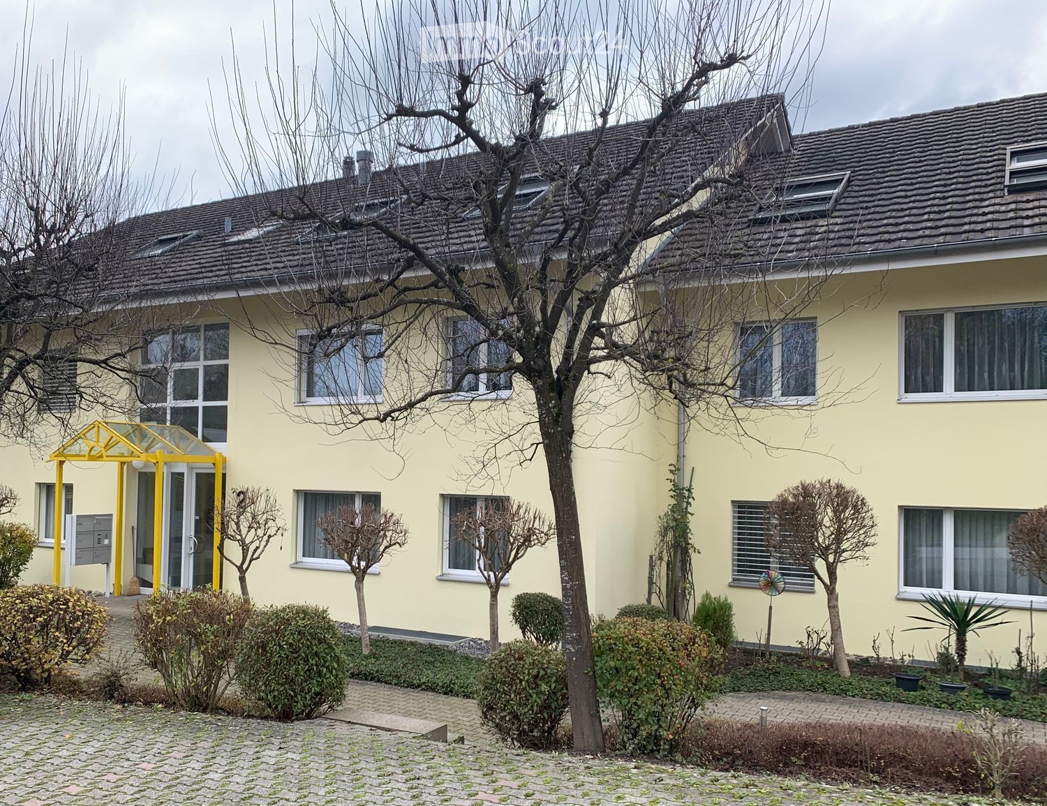 Yellow building, multiple windows, brick pavement, trees, bushes, mailbox