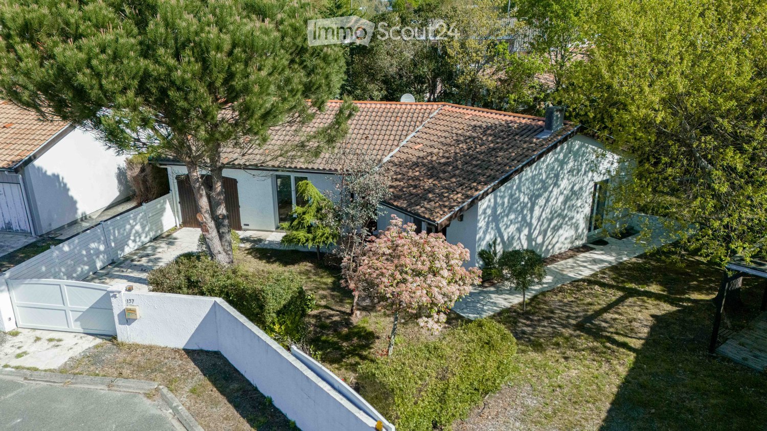 Aerial view of a two-story house with a tiled roof, surrounded by trees and a well-maintained garden. The house has a white exterior with multiple windows and a small porch area.