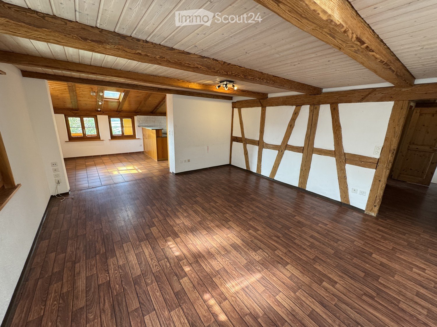 Empty room with wooden flooring, exposed wooden beams, white walls, and a small kitchen area visible in the background.