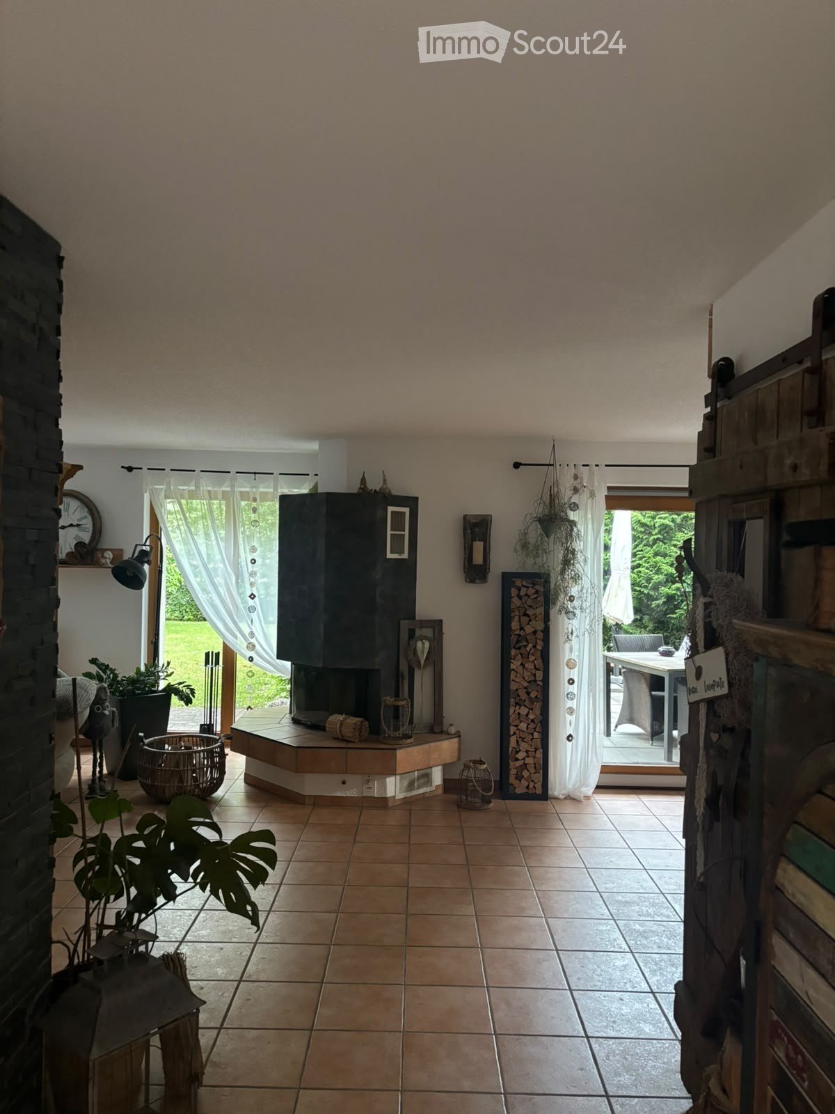 Bright living room with tiled floor, fireplace, large windows with curtains, and outdoor seating area visible through the glass doors.