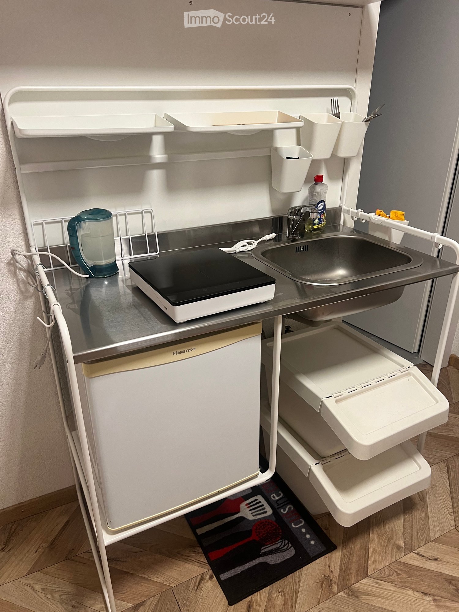 Compact kitchen with a sink, faucet, induction cooktop, electric kettle, refrigerator, and dish rack. Includes a trash bin and a small mat with kitchen utensil graphics.