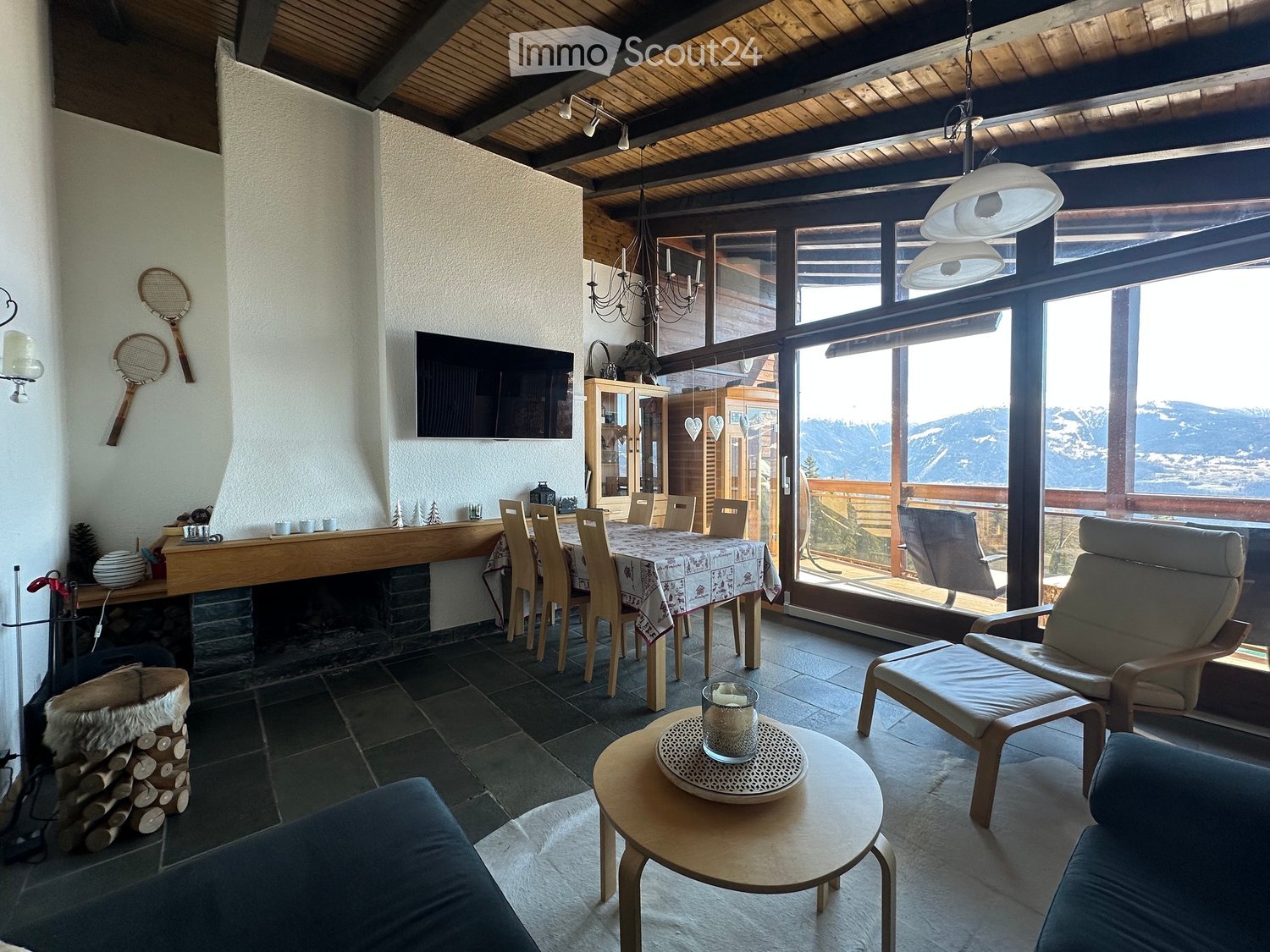 Bright living room with large windows offering a scenic mountain view, featuring a sofa, chairs, a coffee table, a dining area with a table and chairs, a fireplace, and a television mounted on the wall.