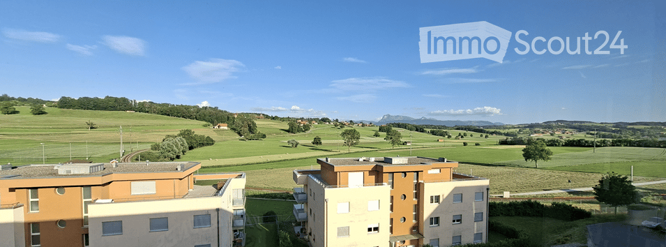 Modern apartment complex with orange and beige facades, surrounded by green fields and distant mountains under a clear blue sky.