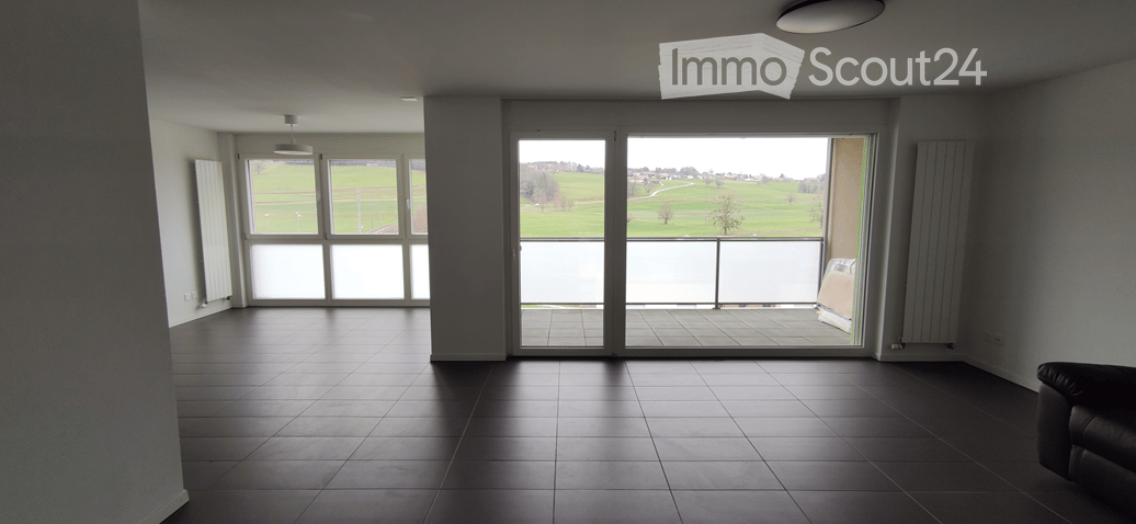 Empty room with large windows and a sliding glass door leading to a balcony, overlooking a green field.