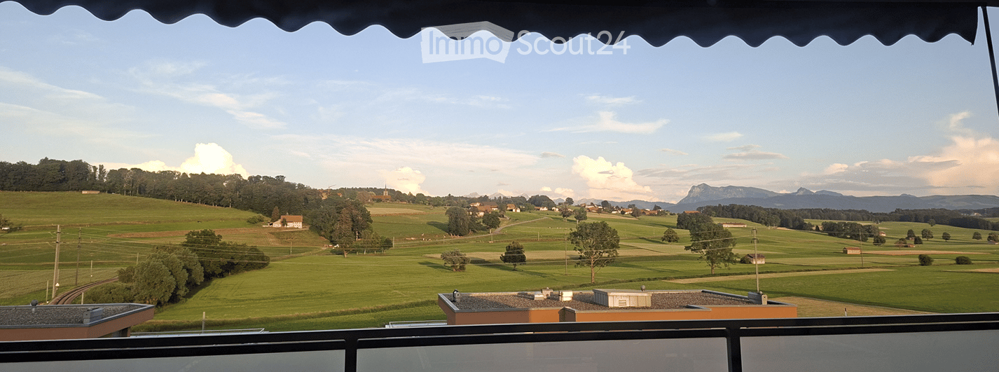 Balcony view overlooking a green, hilly landscape with scattered trees, fields, and distant mountains under a partly cloudy sky.