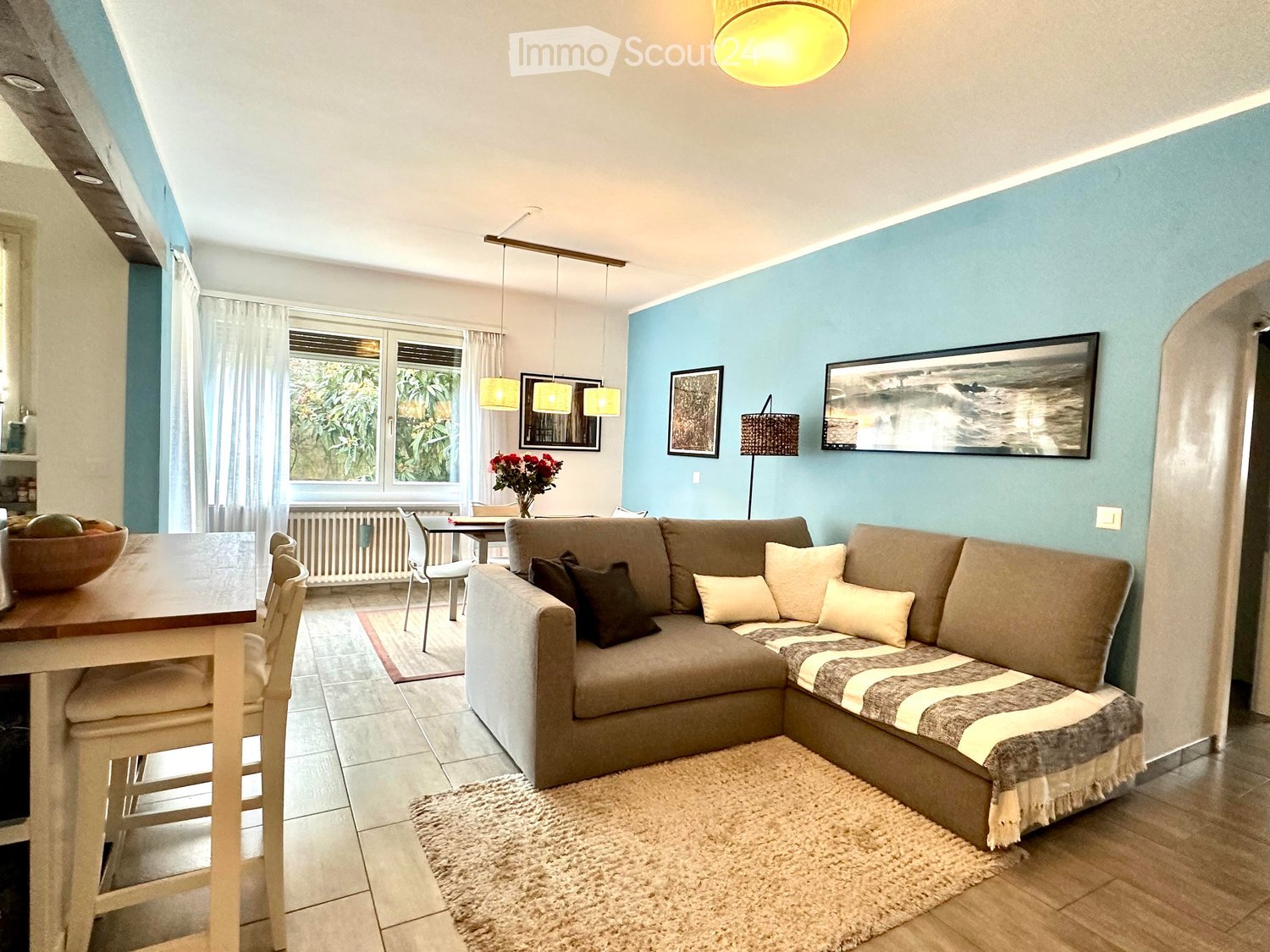 Modern living room with a grey sectional sofa, coffee table, dining table, chairs, and decorative items including a flower vase and picture frames.