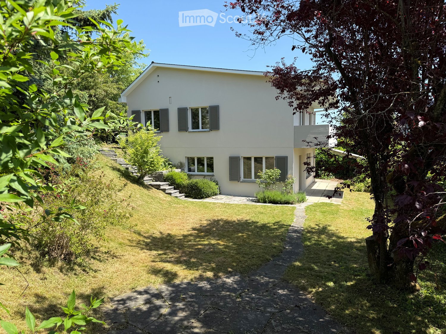 A two-story white house with multiple windows and green shutters, surrounded by a well-maintained garden with a stone pathway leading to the entrance.