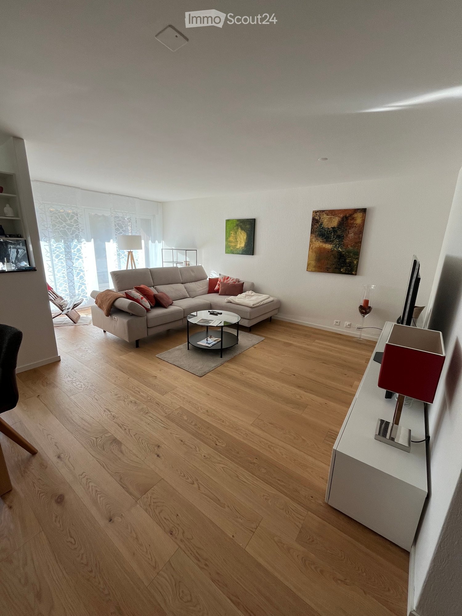 Modern living room with light wooden flooring, grey sectional sofa, coffee table, and large windows with sheer curtains.