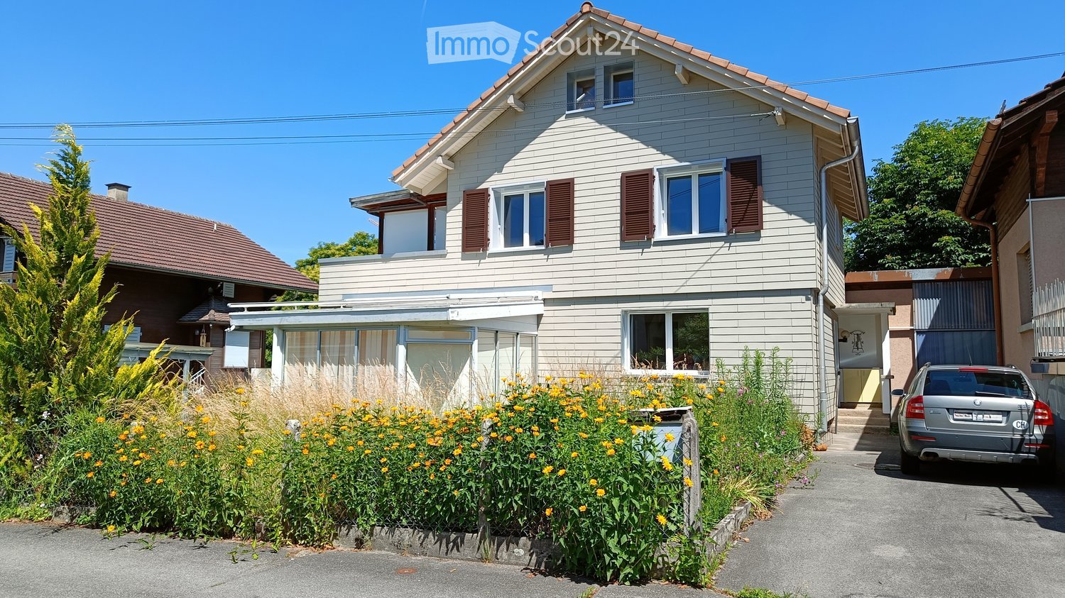 Two-story house with white siding, red-brown shutters, and a small balcony. The house has a well-maintained garden with yellow flowers and a parked car in the driveway.