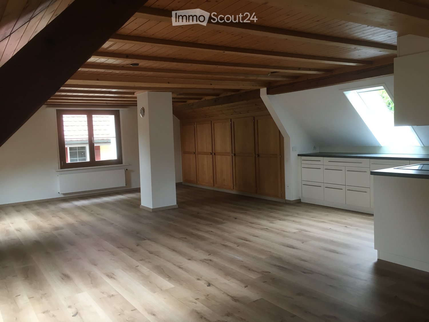 A cozy attic with wooden floor, white ceiling, white cabinets, large window with wooden shutters, built-in wardrobe, ceiling with wooden rafters, skylight
