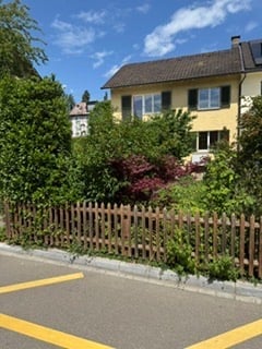 Two-story house with yellow walls, dark roof, multiple windows, surrounded by greenery and a wooden fence.