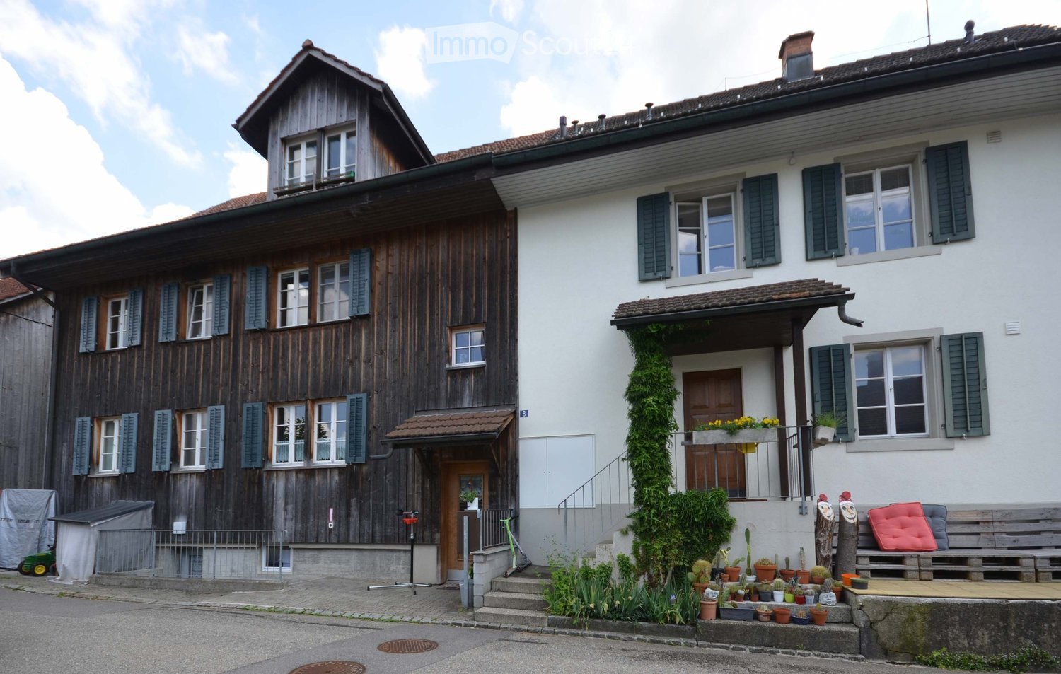 Two-story house with a wooden facade on the left and a white facade on the right, featuring multiple windows with shutters, a small porch, and a garden area with potted plants and a bench.