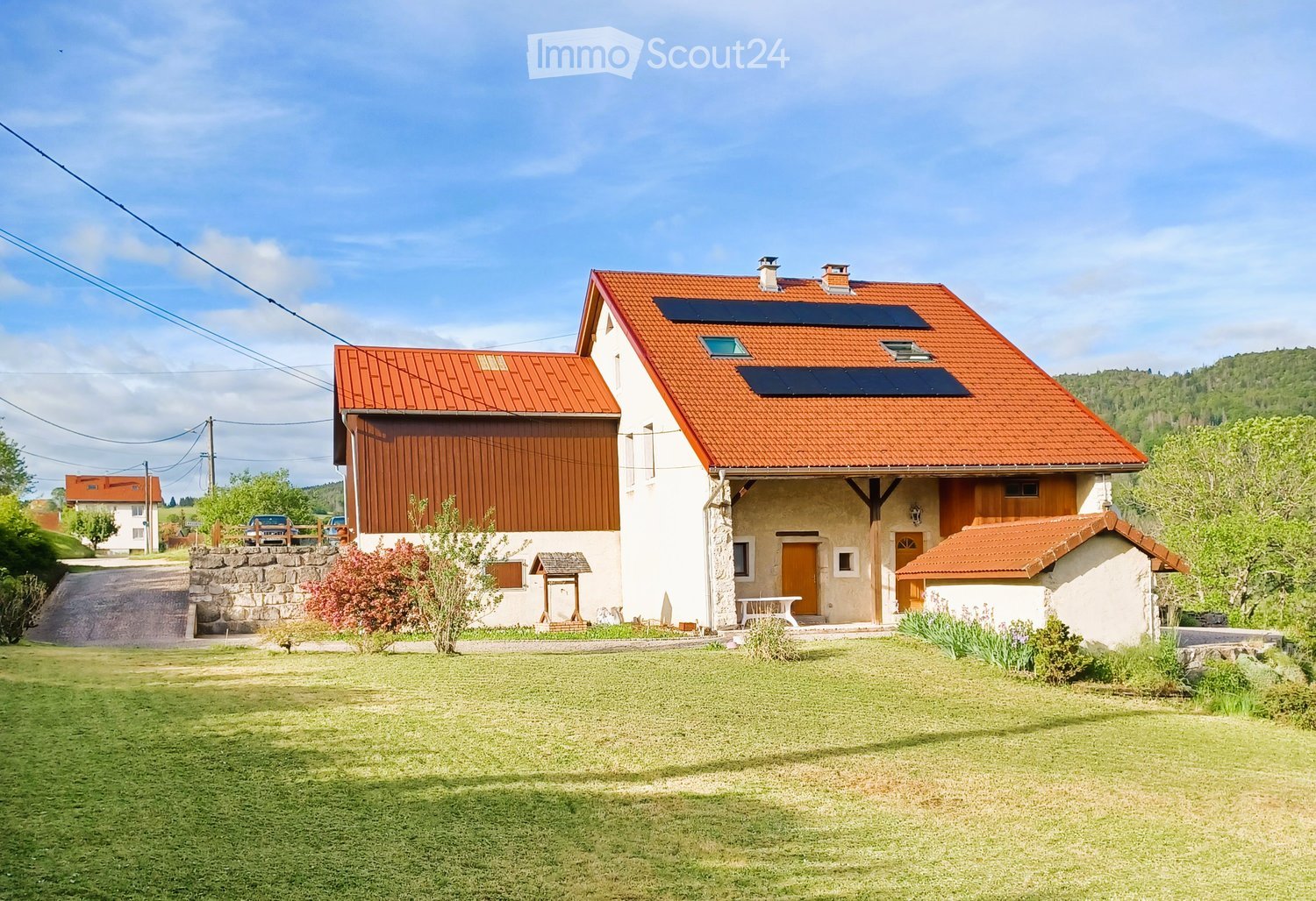 Two-story house with red-tiled roof, solar panels, stone and wood exterior, surrounded by lawn and trees.