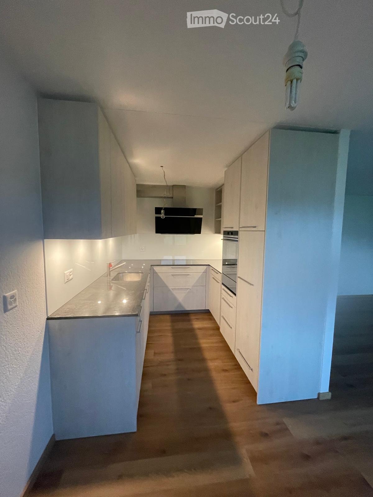 Modern kitchen with white cabinets, wooden floor, sink, and oven. Minimalist design with clean lines and bright lighting.