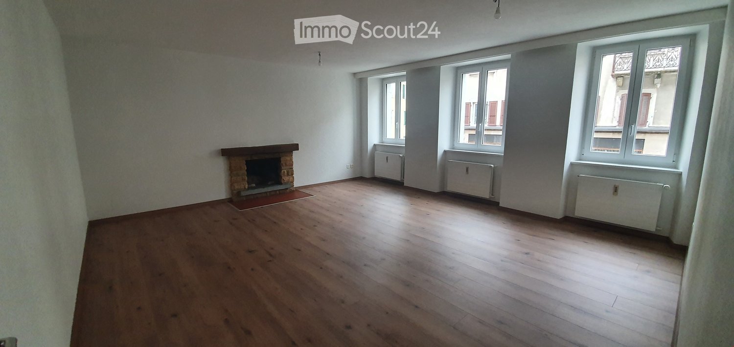 Empty room with wooden flooring, white walls, large windows, and a fireplace with a red rug in front.