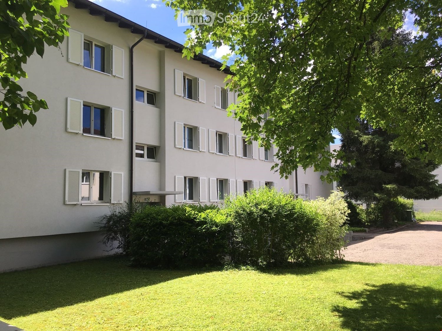 A two-story white building with multiple windows and shutters, surrounded by greenery and a well-maintained lawn.