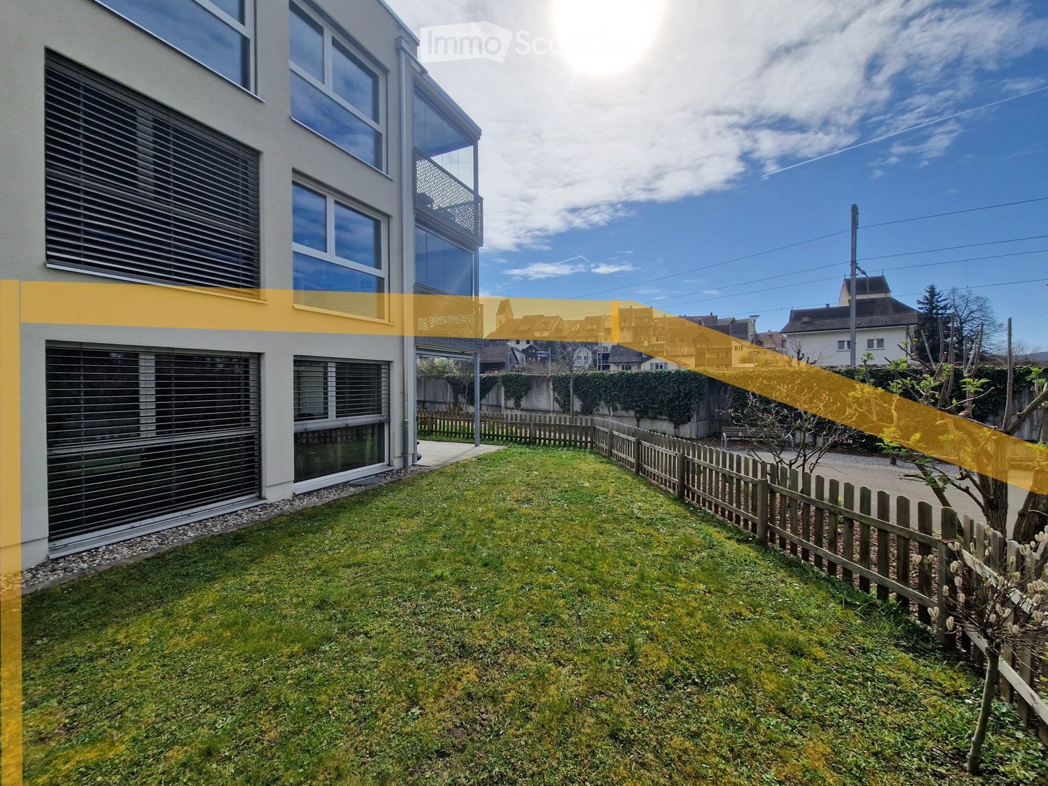 Well-maintained lawn with wooden fence, small tree, and modern house with large windows and balcony in the background.