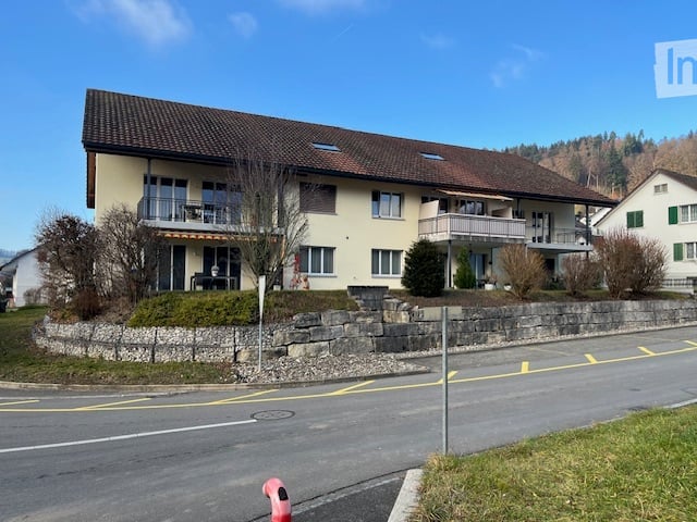 Two-story residential building with balconies, stone retaining wall, and surrounding greenery.