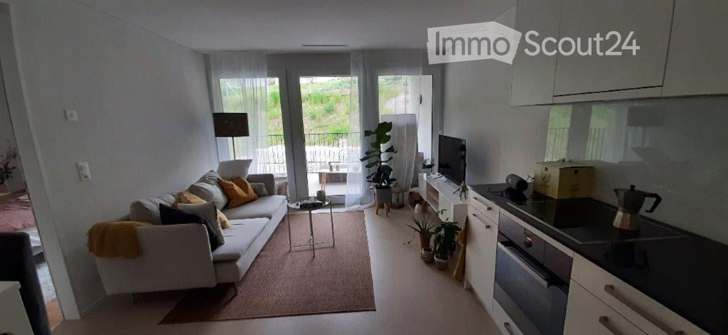 Open-plan living and dining area with a modern sofa, coffee table, and kitchen area featuring an oven, stove, and fridge. Large windows and a balcony are visible.