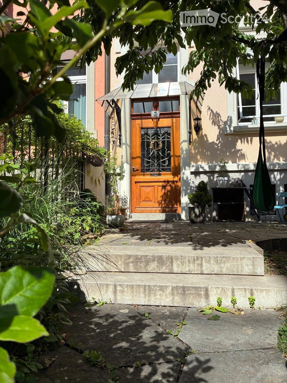 Front entrance of a house with a wooden door, small porch, and surrounding greenery.