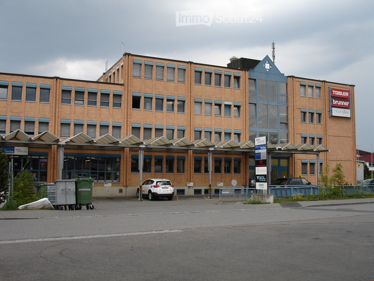 multi-story office building, multiple windows, large car park, cars parked in front, sign for PanGas Marché