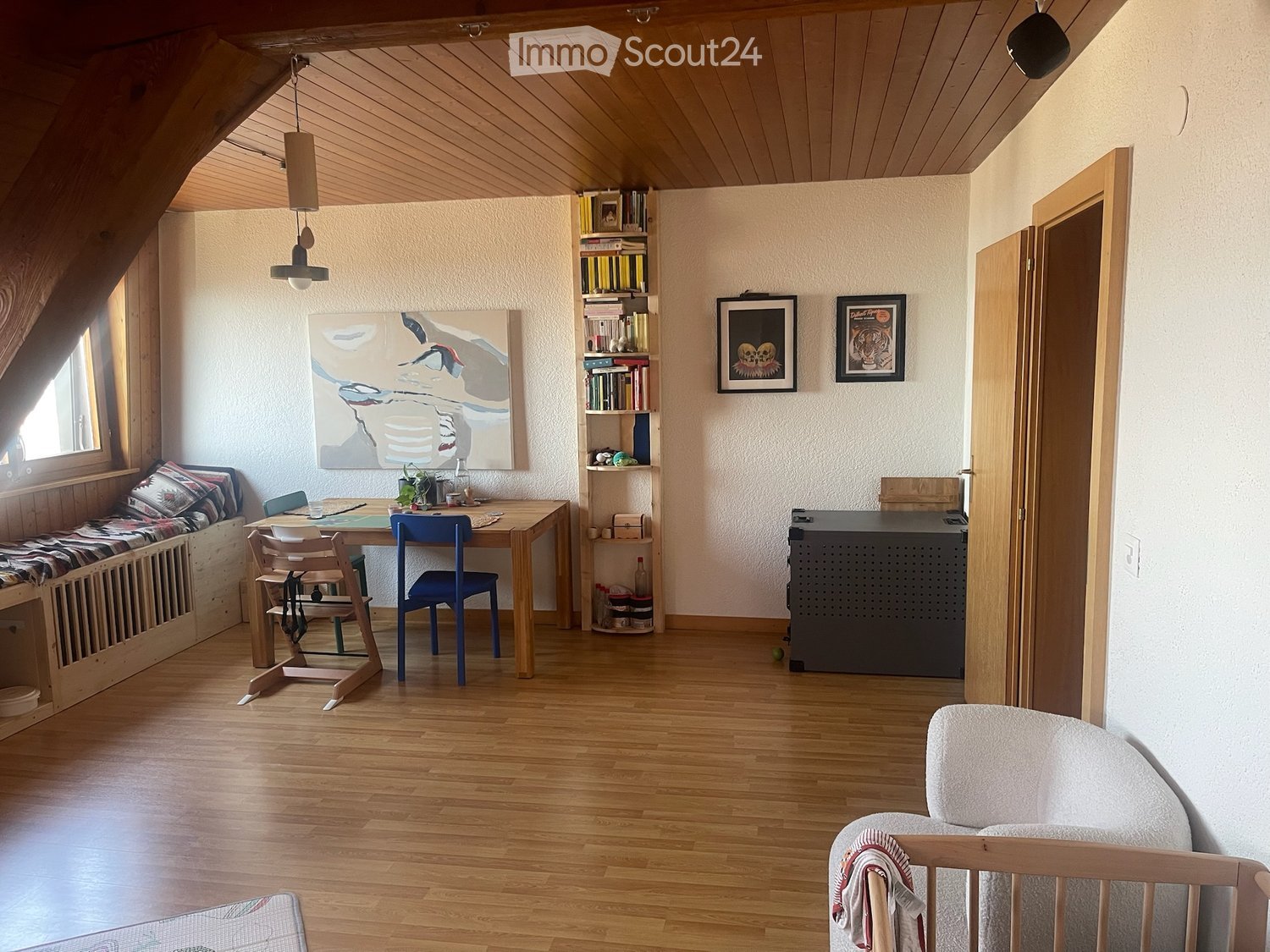 Cozy attic room with wooden floors, slanted ceilings, and large windows. Features a built-in bench seating area, a wooden dining table with chairs, a bookshelf, and a small cabinet.