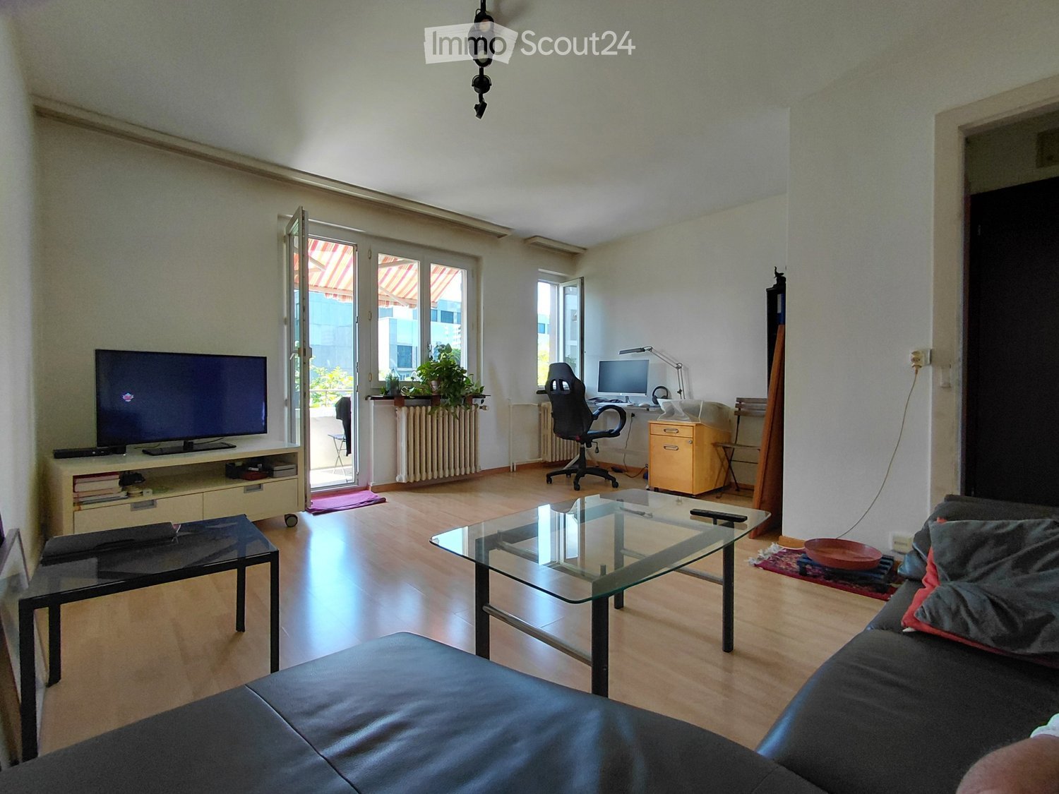 Living room with a sofa, coffee table, TV stand, desk with computer, and a glass table. The room has wooden flooring and large windows with a view of the outside.