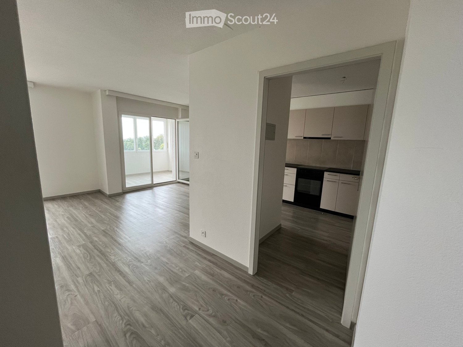 Empty living space with wooden flooring, white walls, and a sliding glass door leading to a balcony. A small kitchen area with cabinets and an oven is visible in the background.