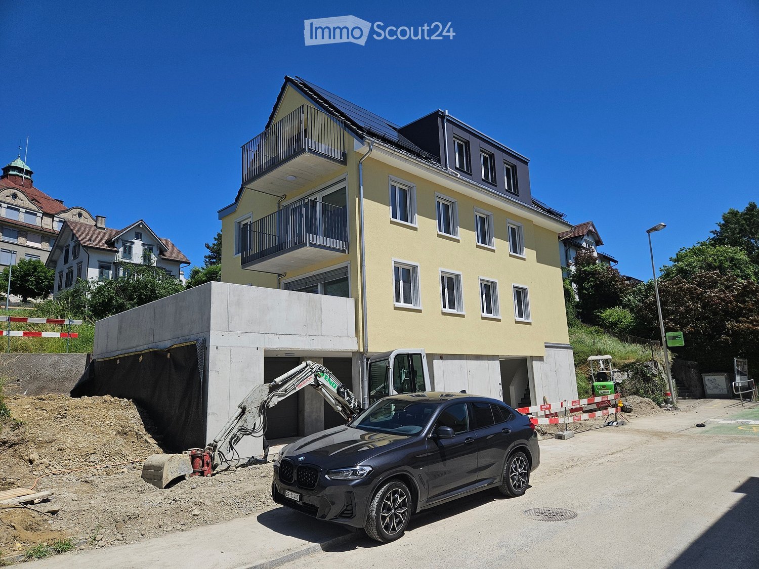 A multi-story residential building with a yellow facade, multiple windows, and balconies. A car is parked in front, and construction work is visible with an excavator and a partially constructed garage.