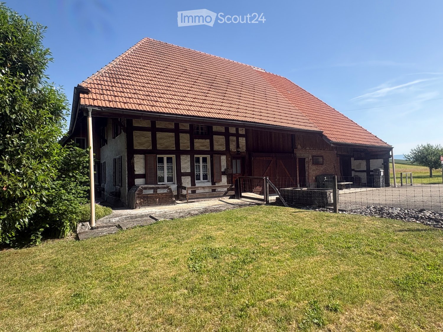 Traditional two-story house with red-tiled roof, wooden beams, and large windows. Includes a wooden bench, fenced area, and grassy lawn.