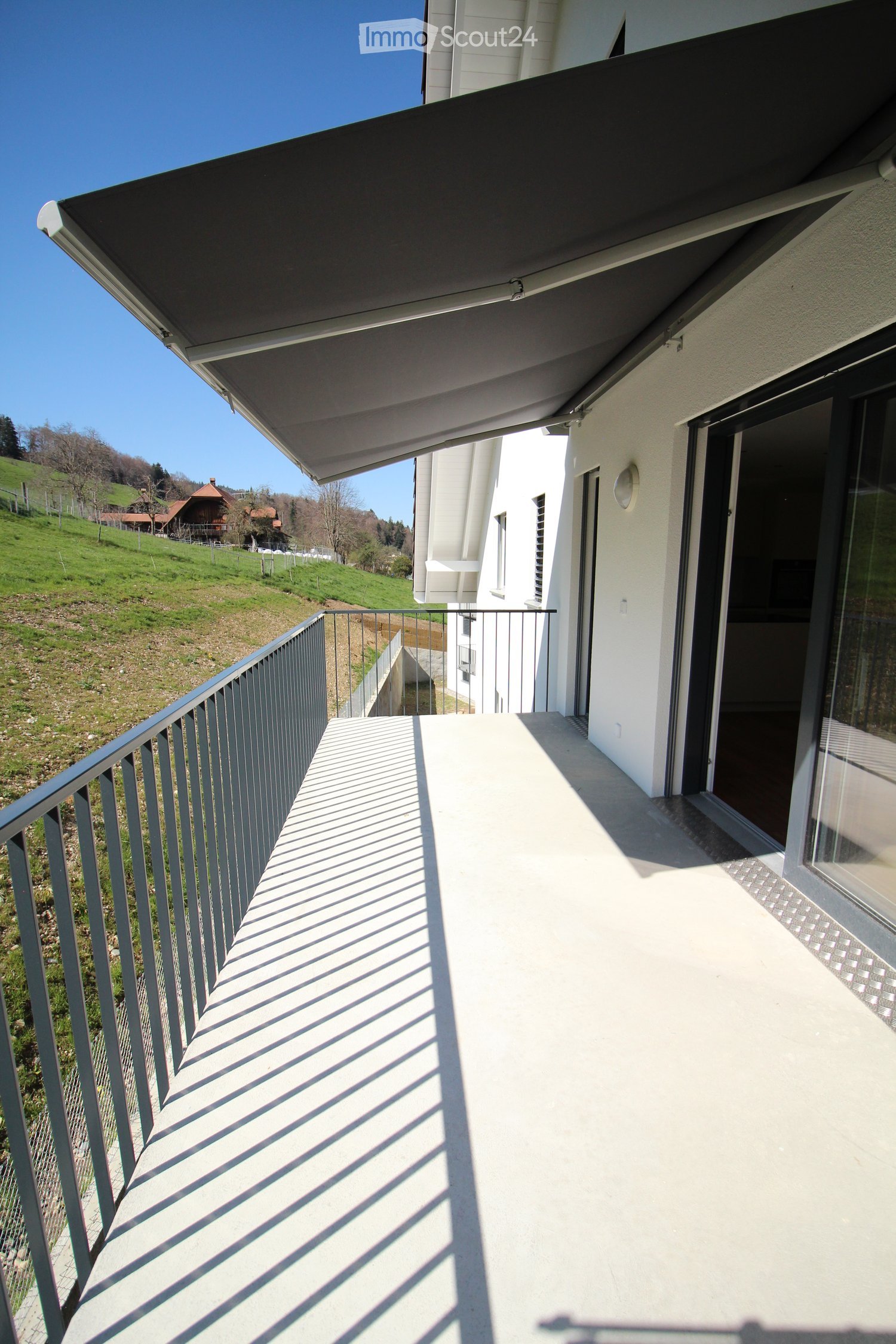 Balcony with a metal railing, covered by a retractable awning, overlooking a green hillside with scattered houses.
