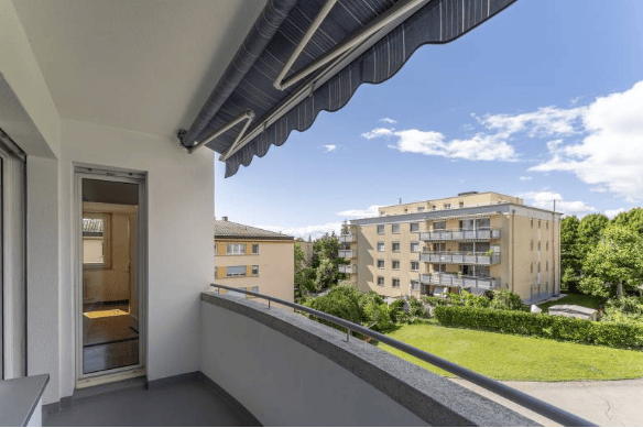 Balcony with a view of a residential area, featuring a metal railing, a sliding door leading to an interior space, and a clear blue sky with scattered clouds.