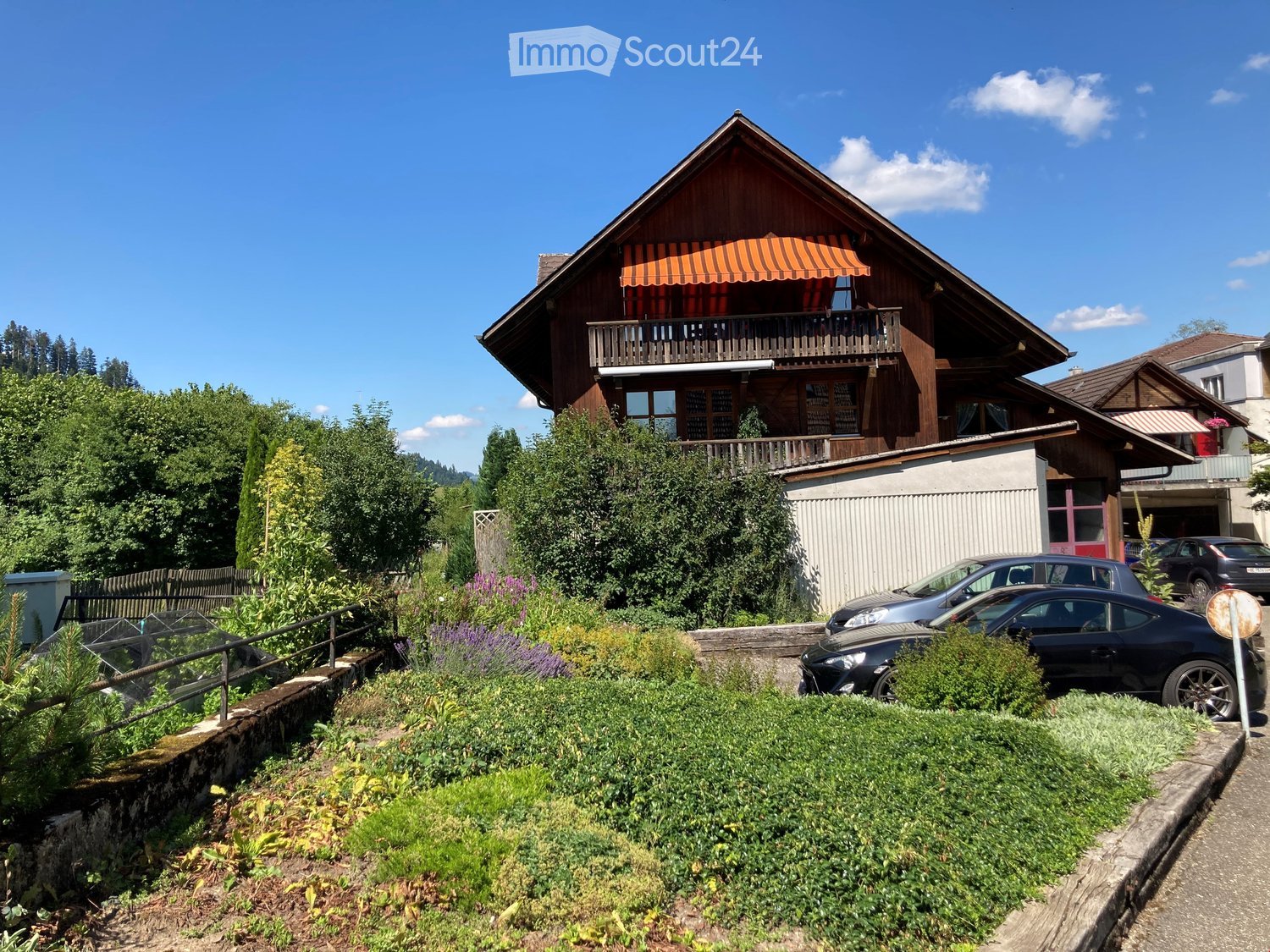 A two-story wooden house with a balcony, surrounded by greenery and parked cars.
