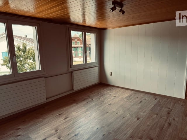 Empty room with wooden flooring, white walls, large windows, and a wooden ceiling.