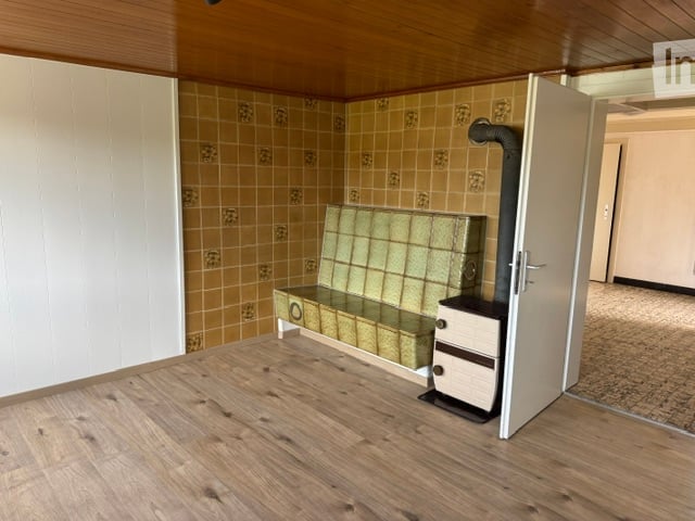 A small, furnished room with a wooden floor, tiled walls, a bench, and a heater. The room has a simple, functional design with a door leading to another area.