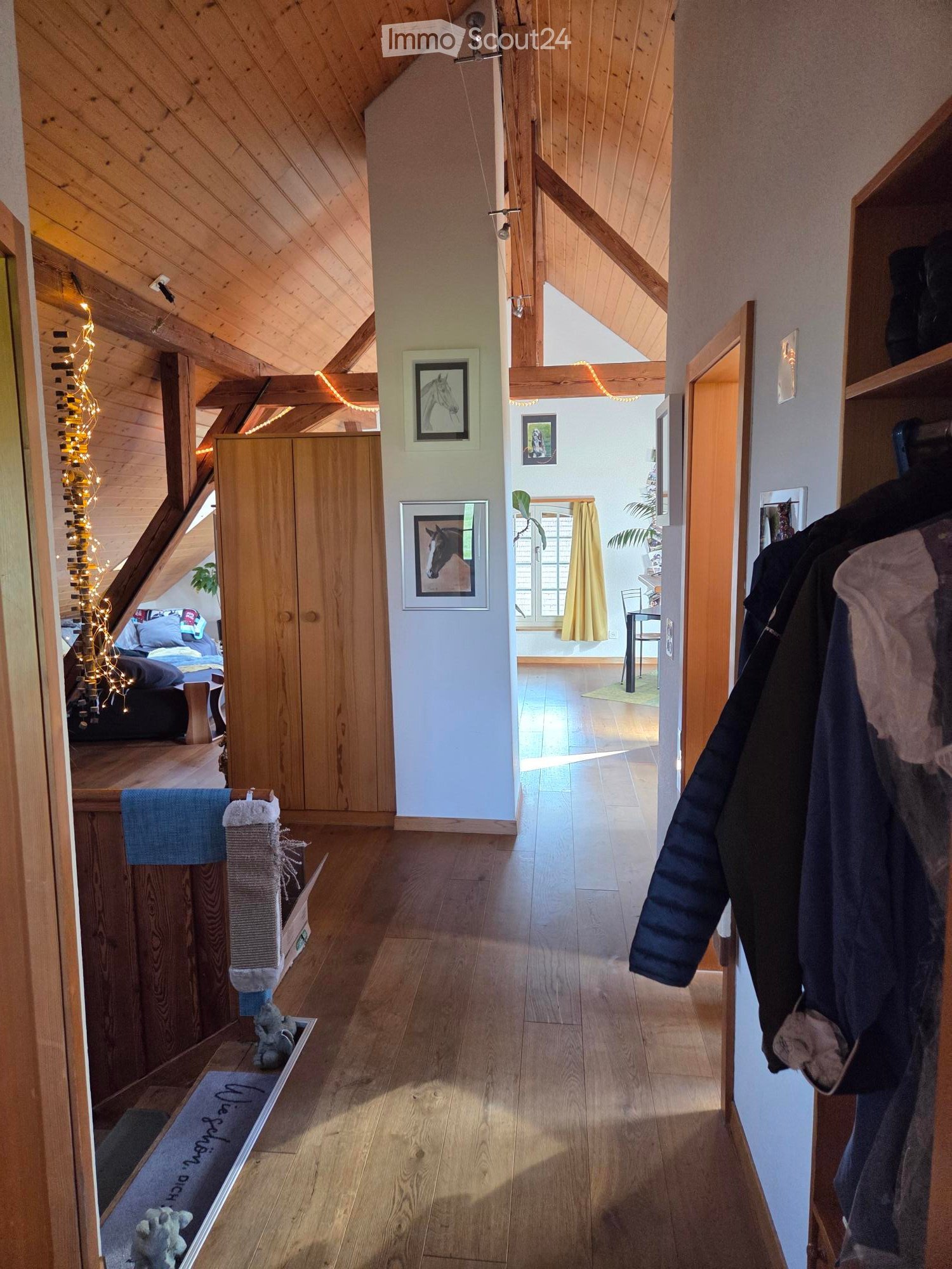 Bright hallway with wooden floors and exposed wooden beams, leading to a bedroom area with a bed and a living area with a chair and table.