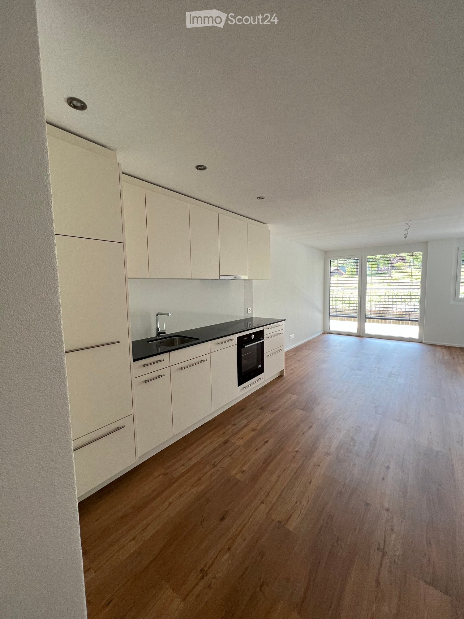 Empty kitchen with light-colored cabinets, a sink, and a built-in oven. The floor is made of light wood, and there is a large window with blinds leading to an outdoor area.