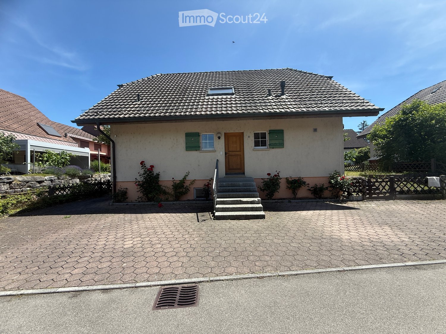 Single-story house with beige siding, green shutters, and a wooden front door. The roof is tiled, and there is a small staircase leading to the entrance. The house is surrounded by a low fence and some flowering plants.