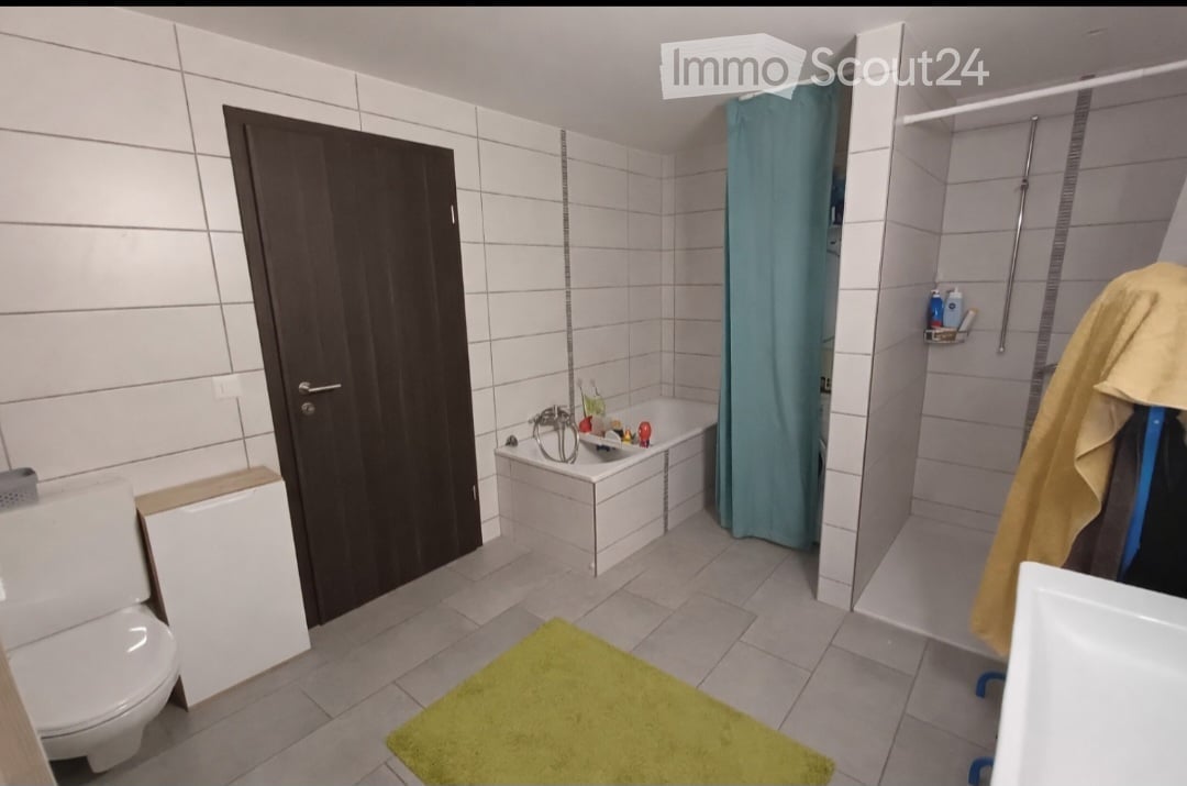 Modern bathroom with white tiled walls and floor, featuring a toilet, bathtub, shower area with a blue curtain, and a yellow bath mat on the floor.