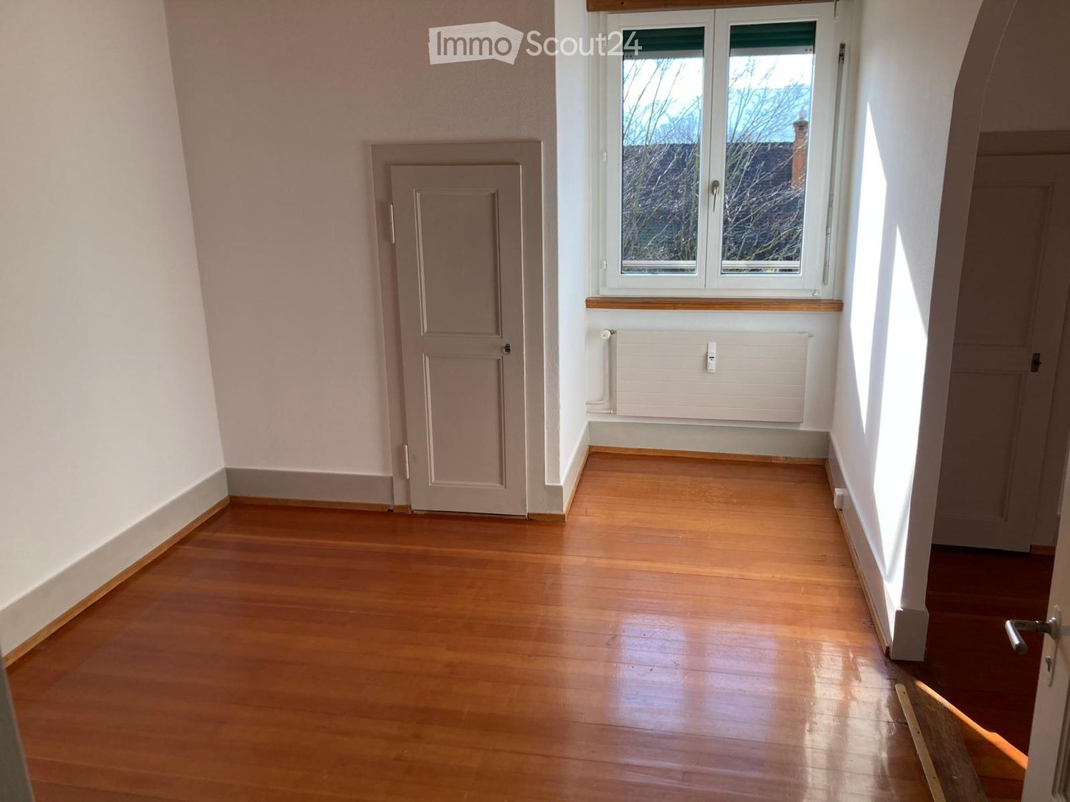 Empty hallway with wooden flooring, white walls, a window with a radiator below, and two doors.