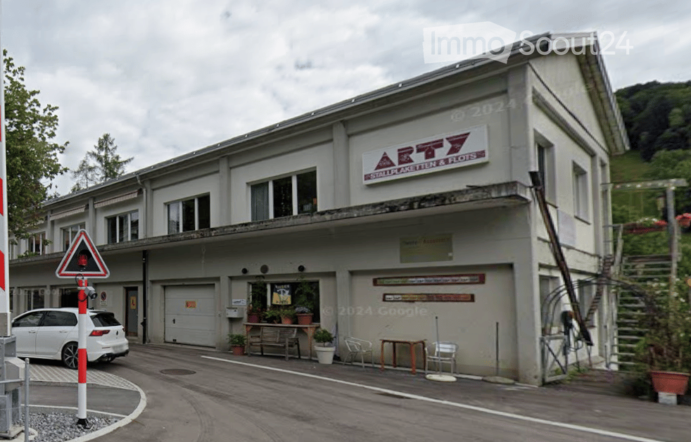 Two-story building with a garage, signage reading "ARTY Stallplaketten & Plots", and outdoor seating area with tables and chairs.