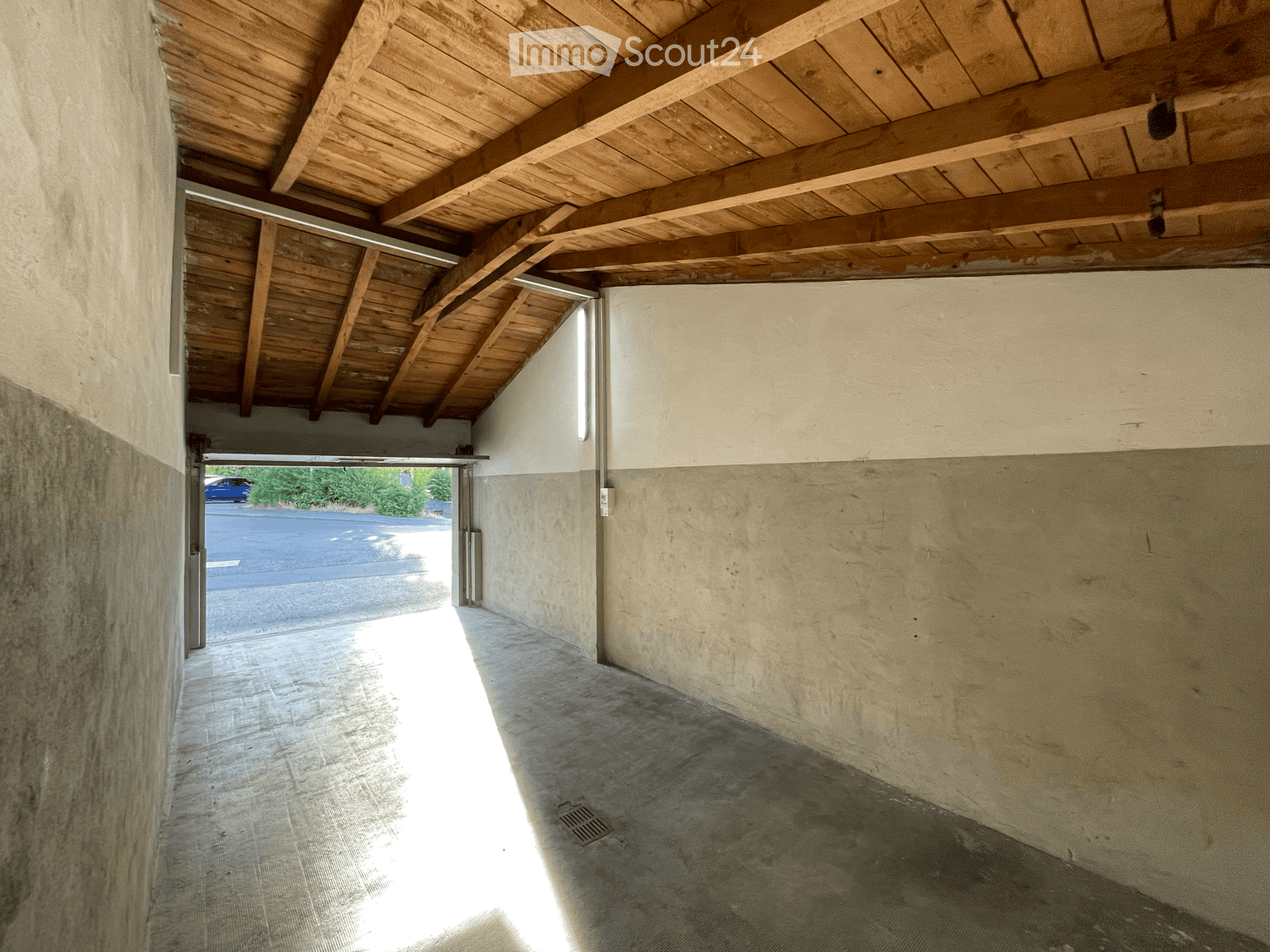 Empty covered corridor with concrete walls and a wooden ceiling, leading to an open garage area.