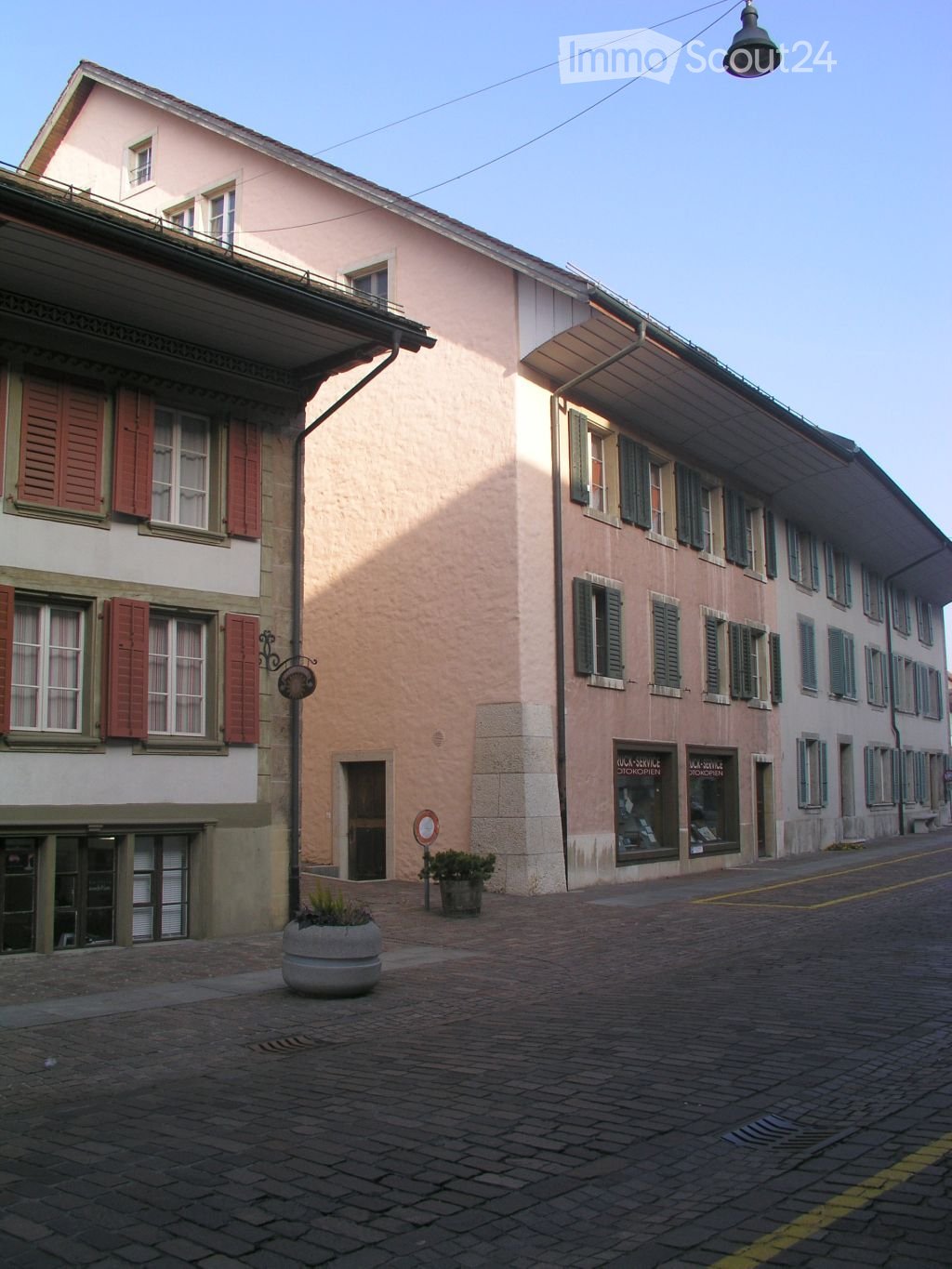Two buildings with multiple windows, red shutters, and a streetlight. The buildings have a brick wall and a stone pavement in front.