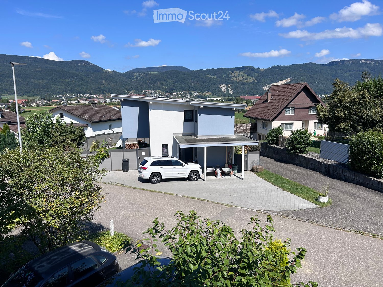 Modern two-story house with a garage, driveway, and surrounding greenery, with mountains in the background.